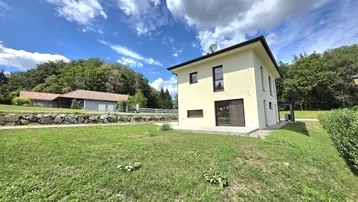 Modernes Einfamilienhaus mit großem Garten und grüner Umgebung unter blauem Himmel.