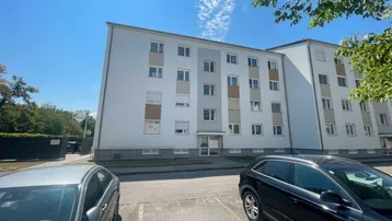 Mehrfamilienhaus mit heller Fassade und Parkplätzen im Freien unter blauem Himmel.