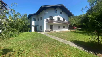 Weißes Einfamilienhaus mit Balkon und großem Rasenbereich, umgeben von grüner Natur.