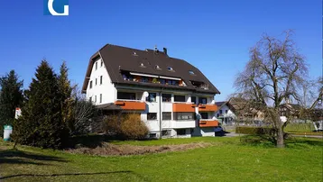 Mehrfamilienhaus mit mehreren Balkonen und Satteldach in grüner Umgebung unter blauem Himmel.
