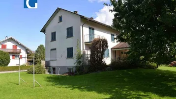 Weißes Einfamilienhaus mit grünem Garten, Balkonen und blauen Fensterläden unter blauem Himmel.
