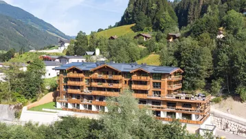 Modernes Gebäude mit Holzfassade und Balkonen, eingebettet in eine grüne Berglandschaft unter blauem Himmel.