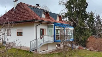 Freistehendes Haus mit rotem Ziegeldach und einem verglasten Wintergarten, umgeben von Bäumen.