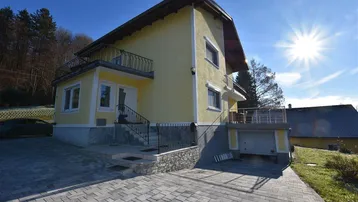 Gelbes Einfamilienhaus mit gepflasterter Auffahrt, Balkonen und integrierter Garage unter strahlendem Sonnenschein.