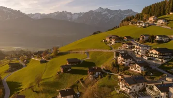 Idyllische Wohnsiedlung auf einem sonnigen Berghang mit atemberaubendem Panorama der umliegenden Alpenlandschaft.