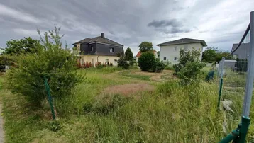 Unbebautes Grundstück mit hohem Gras und Blick auf umliegende Wohnhäuser unter bewölktem Himmel.