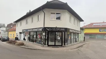 Eckgebäude mit Geschäftsräumen im Erdgeschoss und Wohnungen darüber, an einer Straße gelegen.