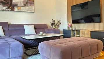 Gemütliches Wohnzimmer mit lila Ecksofa, Couchtisch und modernem TV-Bereich mit Holzverkleidung.