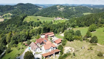 Luftaufnahme eines weitläufigen Anwesens mit mehreren Gebäuden, umgeben von grünen Hügeln und Wäldern.
