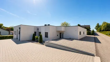 Modernes Einfamilienhaus mit weißer Fassade, Flachdach und gepflasterter Auffahrt unter strahlendem Sonnenschein.