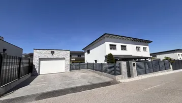 Modernes weißes Einfamilienhaus mit separater Garage, gepflasterter Einfahrt und Zaun.