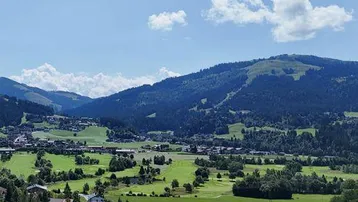 Panoramablick über eine grüne Landschaft mit sanften Hügeln, Bergen und einem Golfplatz unter blauem Himmel.