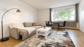Gemütlicher Wohn- und Essbereich mit beigem Ecksofa, Stehlampe und Blick ins Grüne.