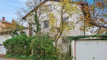 Weißes Einfamilienhaus mit grüner Bepflanzung und Garagenzufahrt in ländlicher Umgebung.