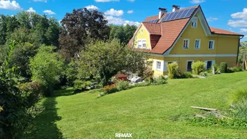 Weitläufiger Garten mit gelbem Wohnhaus, roten Dachziegeln und Solaranlage unter blauem Himmel.
