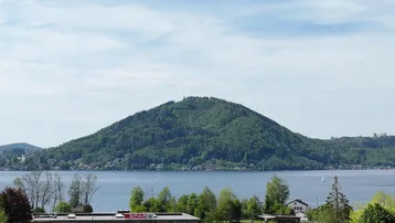 Panoramablick auf den See mit bewaldeten Bergen im Hintergrund und einem Supermarkt im Vordergrund.