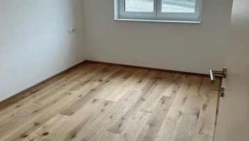 Leeres Zimmer mit Holzboden und großem Fenster, das viel Tageslicht hereinlässt.