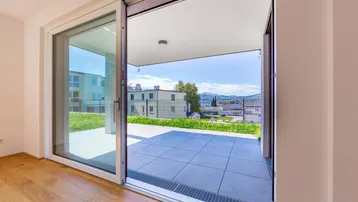 Heller Innenraum mit Holzboden und großen Schiebetüren, die zu einer gefliesten Terrasse mit Bergblick führen.