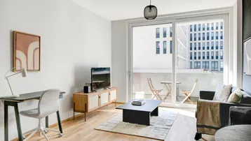 Helles Wohnzimmer mit Holzboden, Schreibtisch und Zugang zu einem Balkon mit Stadtblick.