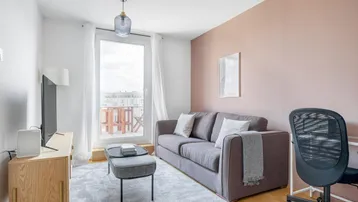 Helles Wohnzimmer mit Sofa, Schreibtisch und Zugang zu einem Balkon, akzentuiert durch eine rosafarbene Wand.