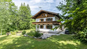Traditionelles Haus im alpenländischen Stil mit Holzbalkon, umgeben von viel Grün und Bäumen.