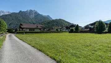 Grünes Feld mit asphaltiertem Weg, umgeben von Häusern und majestätischen Bergen im Hintergrund.