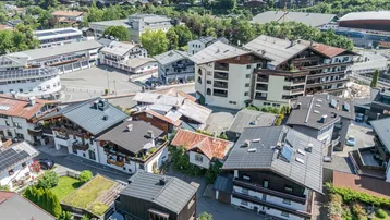 Weitläufige Luftaufnahme einer alpinen Wohngegend mit vielen Häusern, grünen Hügeln und Bergen im Hintergrund.