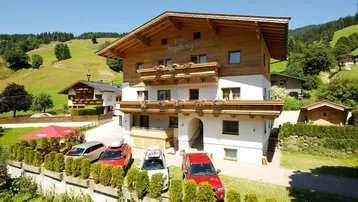 Traditionelles alpines Gebäude mit Holzfassade, blumengeschmückten Balkonen und Parkplätzen vor einer malerischen grünen Hügellandschaft an einem sonnigen Tag.