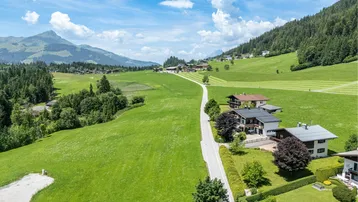 Luftaufnahme einer Wohnsiedlung in einer grünen Berglandschaft mit weiten Wiesen und klarem Himmel.