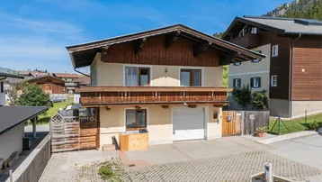 Traditionelles Haus im Alpenstil mit Holzbalkon, Garage und gepflasterter Einfahrt vor Bergkulisse.