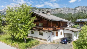 Traditionelles Haus mit Holzfassade und Balkonen, umgeben von grüner Natur und Bergblick.