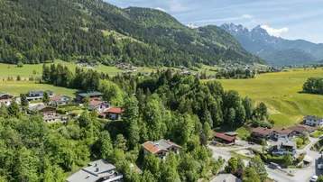 Luftaufnahme eines malerischen Dorfes in den Bergen mit grünen Wäldern und schneebedeckten Gipfeln im Hintergrund.