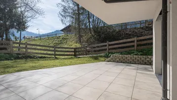 Große geflieste Terrasse mit angrenzender Rasenfläche und Holzzaun, umgeben von grüner Landschaft.