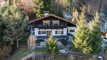 Traditionelles Chalet-Haus in Hanglage mit mehreren Balkonen und umgeben von Bäumen und Schnee.