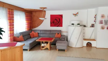 Gemütliches Wohnzimmer mit Ecksofa, traditionellem Kachelofen und Holzelementen an der Decke.
