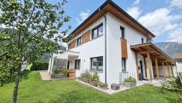 Modernes Einfamilienhaus mit weißer Fassade, Holzdetails und gepflegtem Garten vor Bergkulisse.
