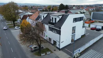 Luftaufnahme eines modernen weißen Gebäudes mit dunklem Dach, umgeben von Wohnhäusern und einer Straße, inklusive Parkplätzen.