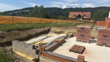 Baustelle mit Fundament und ersten Mauern, im Hintergrund ein Haus mit rotem Dach.