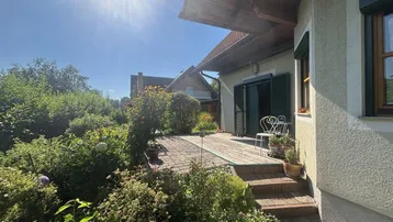 Sonnige Terrasse mit Gartenmöbeln, umgeben von üppigem Grün und Pflanzen.