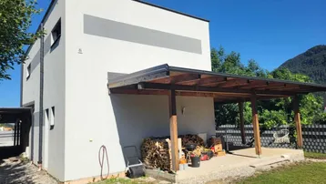 Moderne Hausfassade mit überdachter Terrasse, Holzstapel und Bergblick.