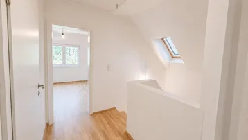 Heller Flur mit Holzfußboden, weiß gestrichenen Wänden, Dachfenster und Zugang zu einem weiteren Raum.