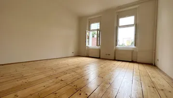 Helles Zimmer mit Holzdielenboden und zwei Fenstern, die viel Tageslicht hereinlassen.