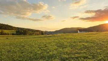 Weitläufiges grünes Feld mit sanften Hügeln und einem Haus in der Ferne bei Sonnenuntergang.
