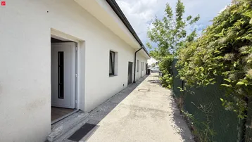 Schmaler Betonweg entlang der weißen Hausfassade mit offener Eingangstür und grüner Bepflanzung.