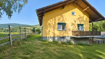 Gelbes Einfamilienhaus mit Holzbalkon und grünen Fensterläden in ländlicher Umgebung.