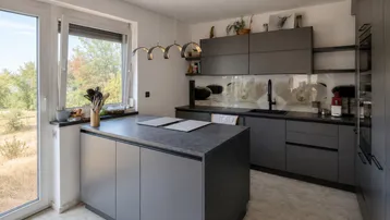 Moderne Küche mit Kücheninsel, grauen Schränken und großem Fenster mit Blick ins Grüne.