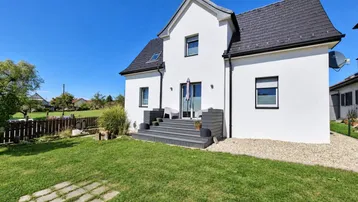 Weißes Einfamilienhaus mit dunklem Dach, kleiner Terrasse und gepflegtem Garten unter blauem Himmel.