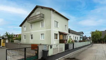 Grünes Wohnhaus mit Balkon, Vorgarten und gepflasterter Zufahrt unter blauem Himmel.