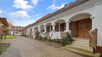Traditionelles Gebäude mit Arkadengang und gepflastertem Hof, umgeben von Bäumen und Sträuchern.