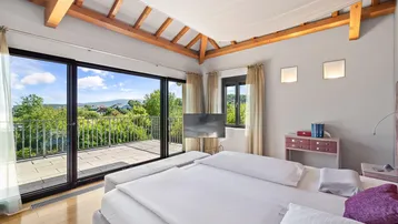 Helles Schlafzimmer mit Holzbalken, Balkonzugang und weitem Blick über die Landschaft.